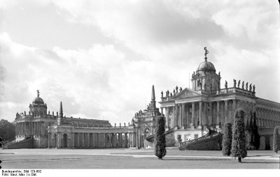 Max Baur (1898–1988), Blick über den Ehrenhof des Neuen Palais auf die Communs, diese sind verbunden durch eine halbkreisförmige Kolonnade die durch zwei Pavillons mit Obelisk eingefasst und in der Mitte mit einem Triumphbogen versehen ist, Schwarz-Weiß-Photographie [o.J.]; Bildquelle: Deutsches Bundesarchiv (German Federal Archive), Bild 170-679, Wikimedia Commons, http://commons.wikimedia.org/wiki/File:Bundesarchiv_Bild_170-679,_Potsdam,_Sanssouci,_Communs.jpg.Creative Commons Attribution-Share Alike 3.0 Germany license.